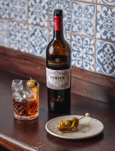 A bottle of Gonzalez Byass Vermouth is placed on a table next to a glass filled with an iced cocktail, garnished with an olive. A small plate holds skewered olives against a backdrop of decorative blue and white tiles.