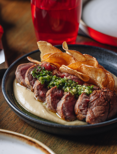 Sliced steak topped with green chimichurri sauce, served on a bed of creamy puree, accompanied by crispy potato chips in a black plate. A glass of red drink is in the background.