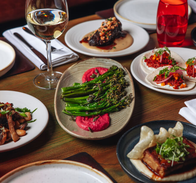 A table set with various dishes including roasted broccolini on a platter with a pink sauce, small plates with red-hued appetizers, crispy pork belly, a glass of white wine, and vibrant decor elements like a red vase.