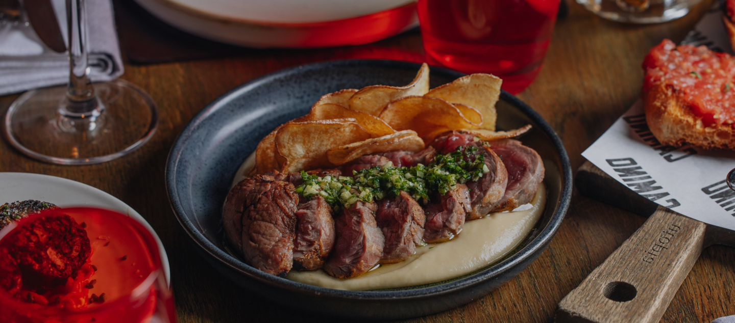 A plate of sliced steak topped with green chimichurri sauce, served with a side of crispy chips on a dark round plate. The table includes drinks, a red sauce in a dish, and a piece of tomato-topped bread.