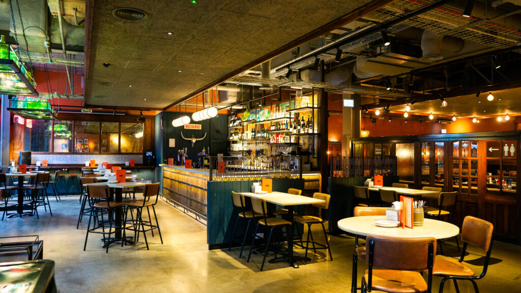 Modern bar interior with high ceilings and industrial design. Features wooden bar with illuminated shelves, high stools, round tables with chairs, and ambient lighting. Warm colors and a relaxed atmosphere dominate the space.