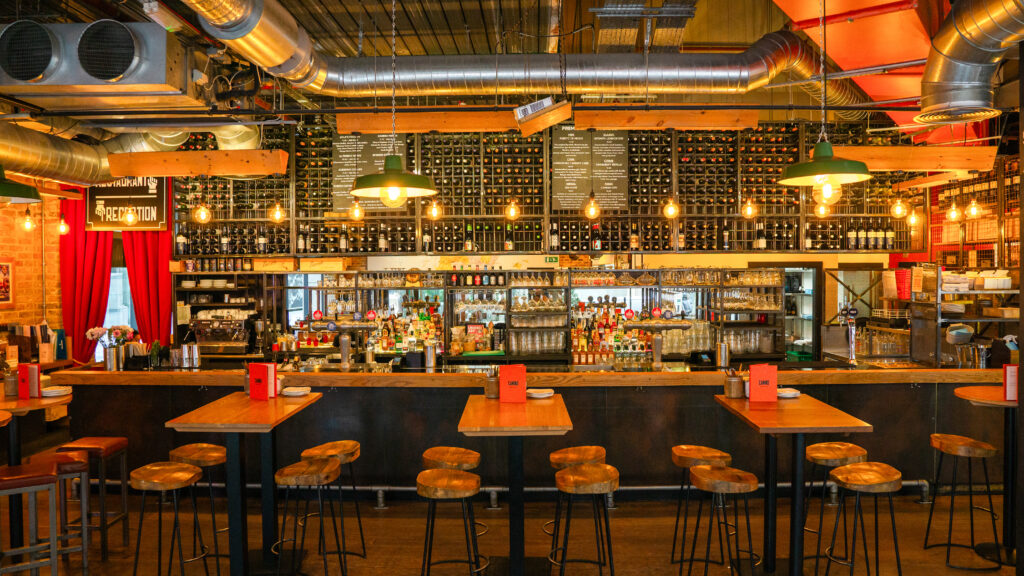 A cozy bar interior with high wooden tables and stools in the foreground. The back wall is lined with shelves of wine bottles and illuminated by warm, hanging lights. Exposed ducts and industrial decor add to the atmosphere.