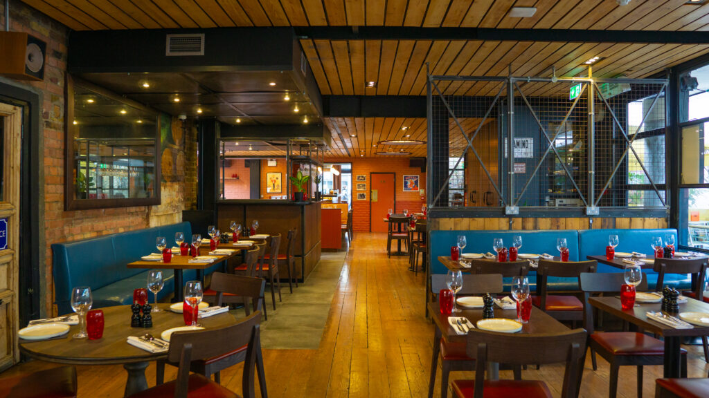 A cozy restaurant interior with wooden floors and ceiling. Tables are set with glasses and napkins, accompanied by red pepper mills. Blue cushioned benches line the walls. Warm lighting and framed pictures create a welcoming atmosphere.