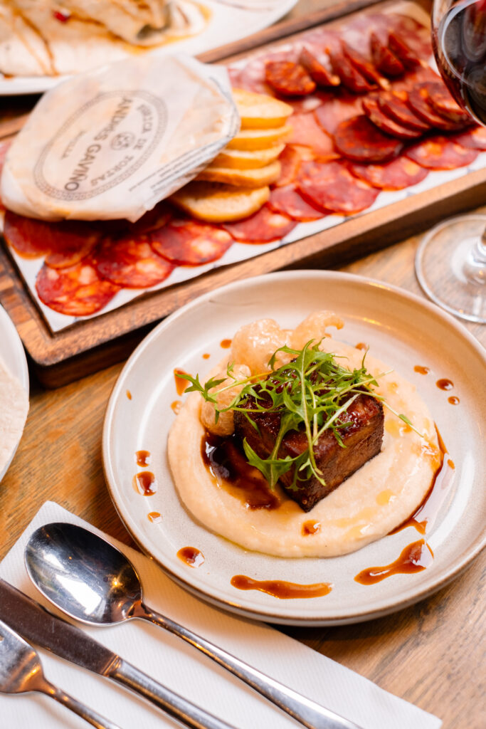 A plate of food featuring a piece of meat garnished with greens, served on a creamy sauce. In the background, a charcuterie board with sliced cured meats and crackers is visible, along with a glass of red wine. Silverware is set beside the plate.