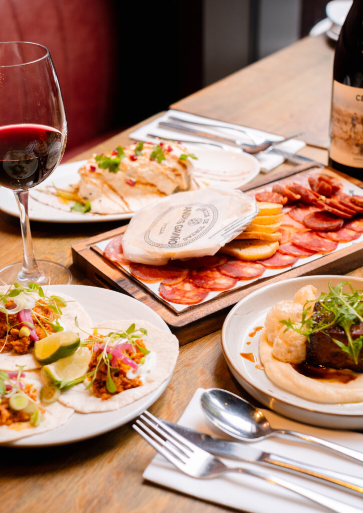 A wooden table set with a glass of red wine, a plate of tacos with lime wedges, a charcuterie board with meats and cheeses, and a dish with hummus and greens. A wine bottle is partially visible in the background.