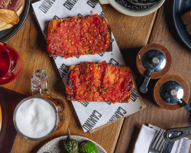 Two slices of toasted bread topped with a tomato mixture sit on a paper-lined wooden board. A glass of beer, salt and pepper shakers, green pepper, and napkin are nearby on a wooden table.