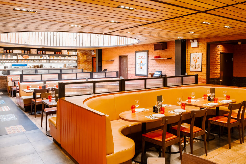 A modern restaurant interior with wooden ceilings and orange U-shaped booth seating. Tables are set with red napkins and glassware. An open kitchen is visible in the background, and the walls are decorated with framed artwork.