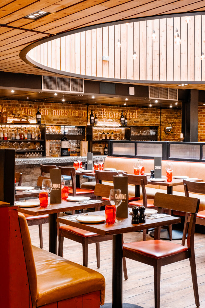 Modern restaurant interior with wooden ceiling and floor. Tables with red candles and menus are neatly arranged. A wine shelf and a welcoming sign in Spanish are visible on the brick wall in the background. Warm lighting creates a cozy atmosphere.