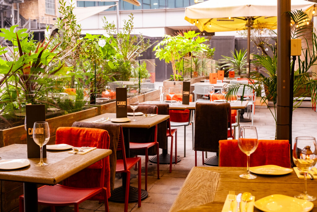 An outdoor restaurant patio with wooden tables and red cushioned chairs, set with wine glasses and napkins. Lush green plants line the area, and a large white umbrella provides shade. The setting appears inviting and peaceful.