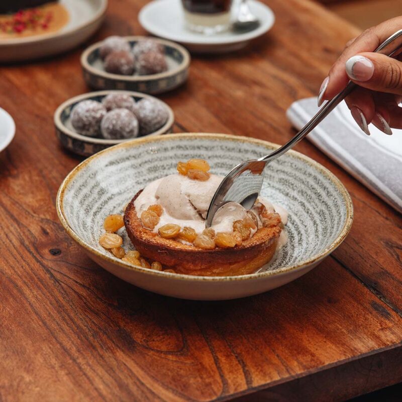 A person scoops a dessert from a bowl with a spoon. The dish contains a pastry topped with ice cream and golden raisins. The bowl is on a wooden table with other small dishes and a cup in the background.