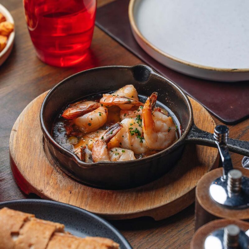 A small cast iron skillet with sizzling shrimp garnished with herbs, set on a wooden board. It sits beside a red glass, plates, and pepper mills on a wooden table.