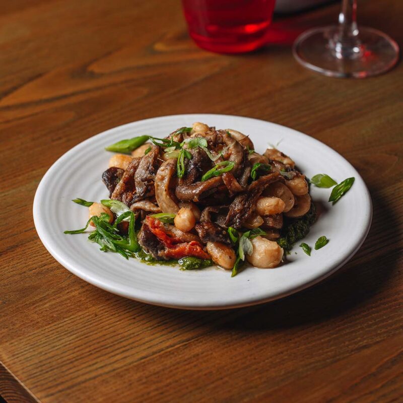 A white plate on a wooden table holds a dish of sautéed mushrooms, white beans, and greens garnished with sliced green onions. There is a glass with a red drink partially visible in the background.