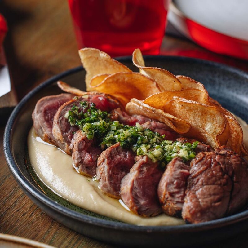 A plate featuring sliced beef with green herbs, served on a light cream sauce, accompanied by a stack of crispy potato chips. A red drink is visible in the background, on a wooden table.