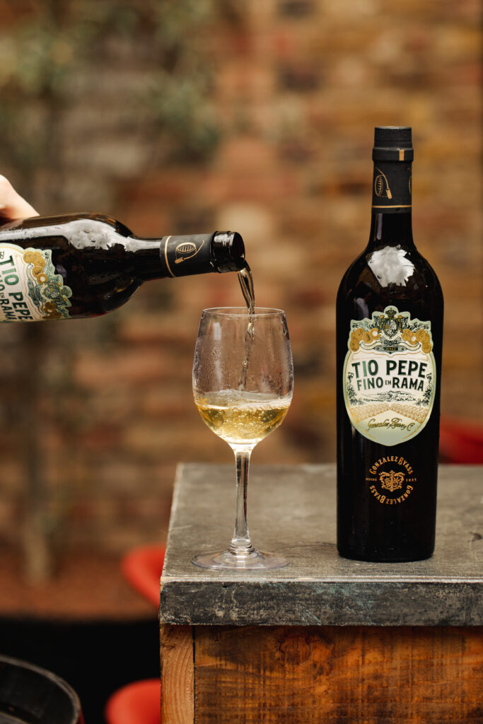 A bottle of Tio Pepe fino sherry is next to a wine glass on a wooden surface. Sherry is being poured into the glass from another bottle. The background features a blurred, rustic brick wall and red accents.