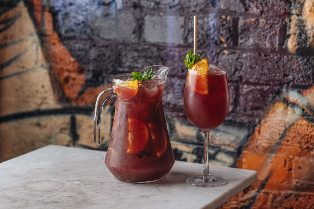 A glass and a pitcher of red sangria sit on a marble table. Both contain slices of orange and are garnished with fresh mint. A colorful graffiti wall serves as the backdrop.