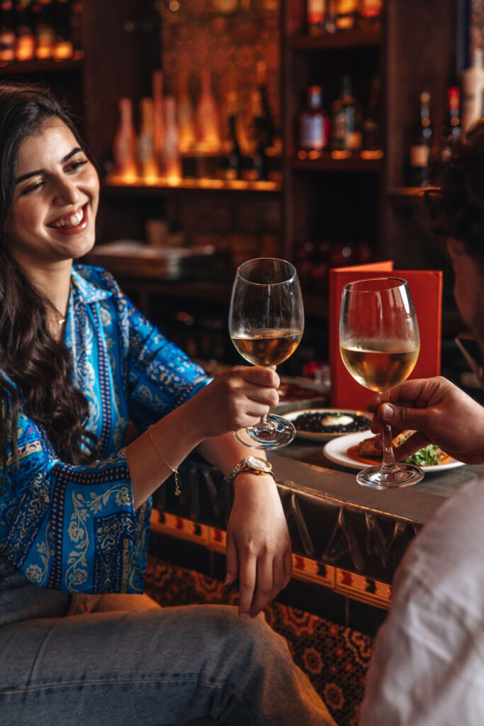 A woman in a blue patterned shirt sits at a bar, smiling and toasting with a glass of white wine. Shelves with bottles are in the background. Another person is partially visible, holding a wine glass. The setting is warmly lit and casual.