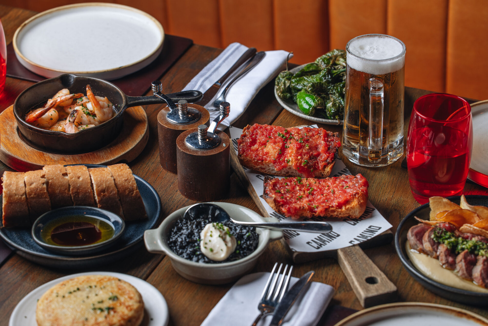 A wooden table is set with various Spanish tapas dishes, including tomato bread, prawns, padrón peppers, a small skillet of food, a beer glass, and a red cup. Plates and silverware are arranged around the colorful dishes.