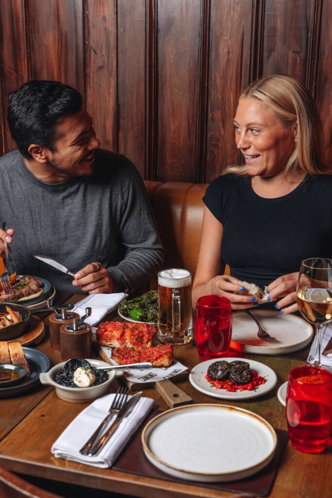 A man and woman sit at a restaurant table, smiling and facing each other. The table is filled with dishes including pizza, a glass of beer, and glasses of wine. The setting has warm lighting and a wooden backdrop.