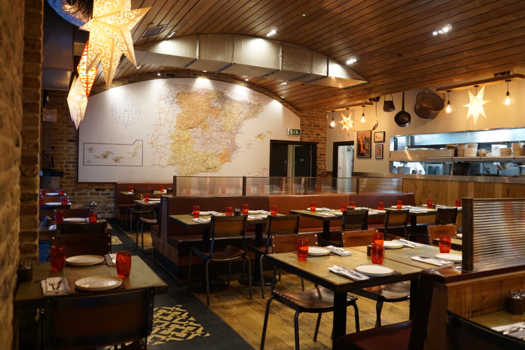 Restaurant interior with wooden ceiling and tables set for dining. Red glasses and white plates are neatly arranged. Decorative star-shaped lights hang overhead. A large map of Spain adorns the back wall near an open kitchen area.