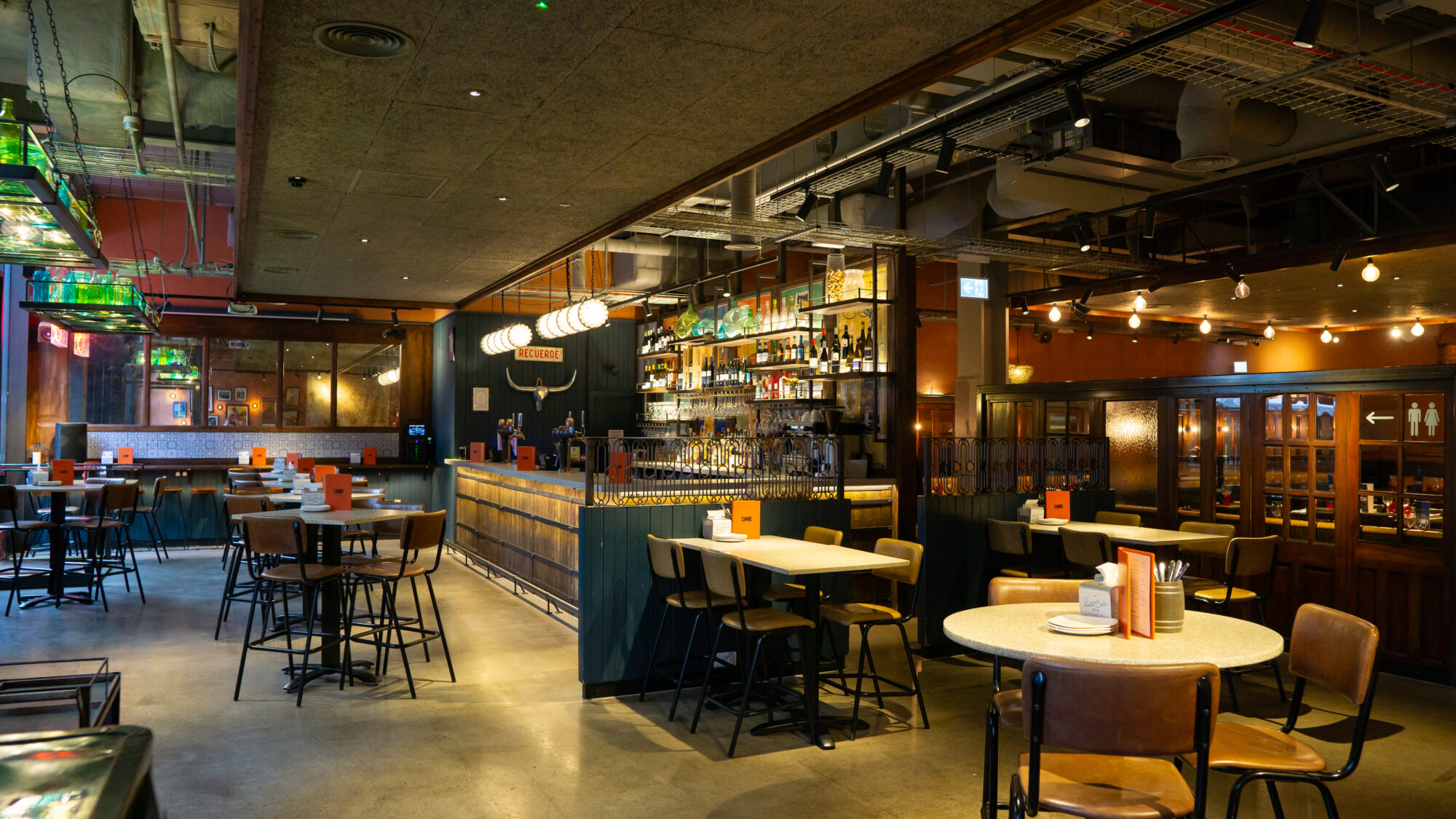 A spacious, modern bar and restaurant with a long counter, high stools, and round tables. The ambient lighting creates a warm atmosphere. Shelves behind the bar display various bottles. The ceiling features exposed pipes and ductwork.