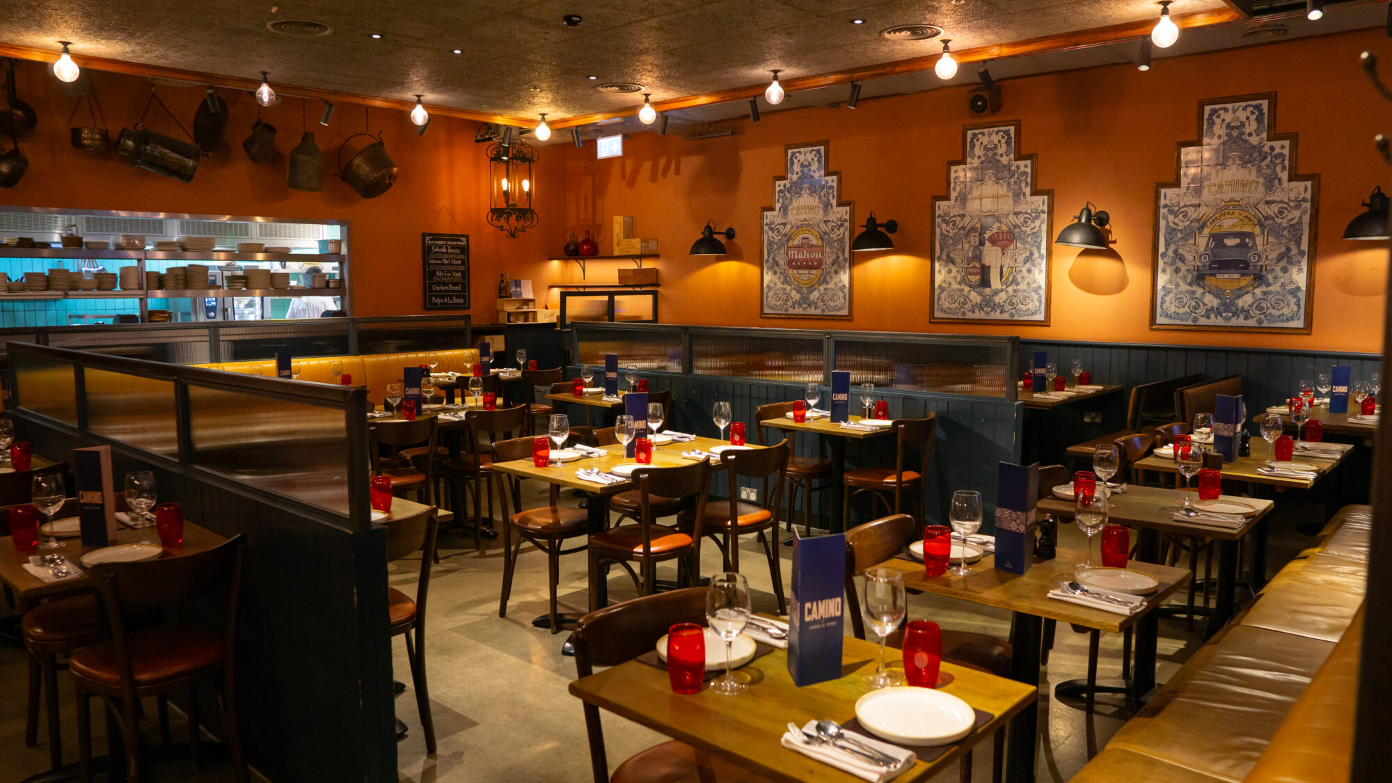 Cozy restaurant interior with wooden tables set for dining, red glass candle holders, and blue menus. Warm lighting and decorative wall art create an inviting atmosphere. Hanging pots and pans adorn walls, and an open view of the kitchen is visible.