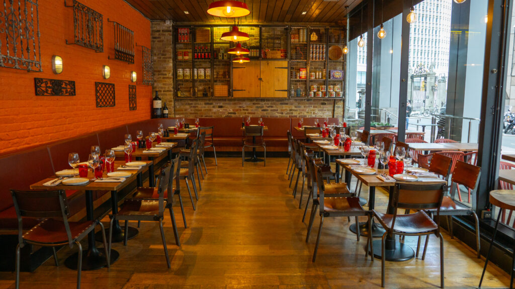 Interior of a cozy restaurant with wooden floors and exposed brick walls. The tables are neatly set with red napkins, glasses, and cutlery. Warm lighting creates an inviting atmosphere, and large windows offer a view of the city outside.