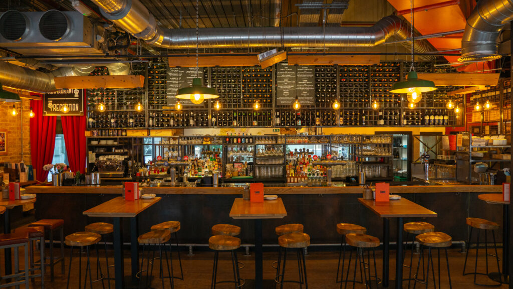 An industrial-style bar interior featuring wooden high tables and stools. The wall behind the bar is lined with wine bottles and glasses. Exposed ductwork and warm pendant lighting add to the atmosphere.