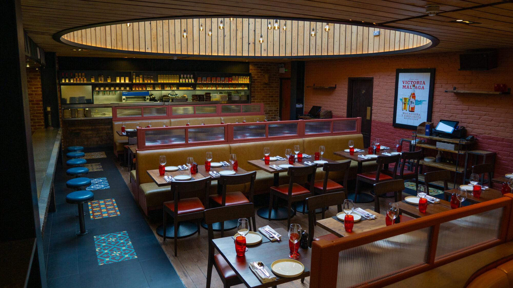 A modern restaurant interior with tables set for dining. Red cushioned benches and chairs surround wooden tables. Blue barstools line a counter. A lit circular ceiling feature enhances the atmosphere, with shelves and kitchen view in the background.