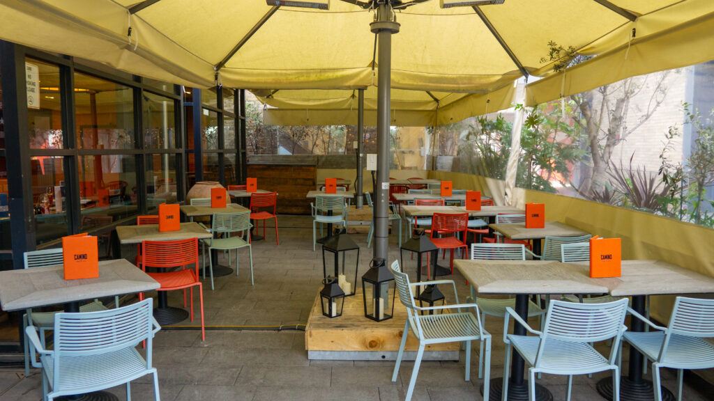 Outdoor cafe with empty tables and multicolored chairs beneath a yellow canopy. Each table has an orange menu labeled 