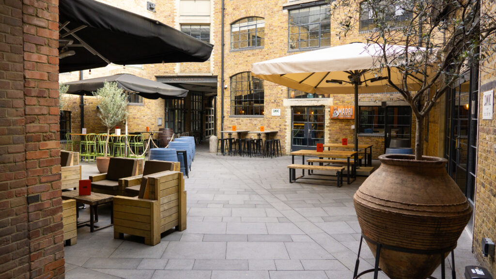 Outdoor patio with brick walls and various seating options, including wooden chairs, bar stools, and a large clay pot plant. Umbrellas provide shade, while industrial-style windows add character to the courtyard setting.