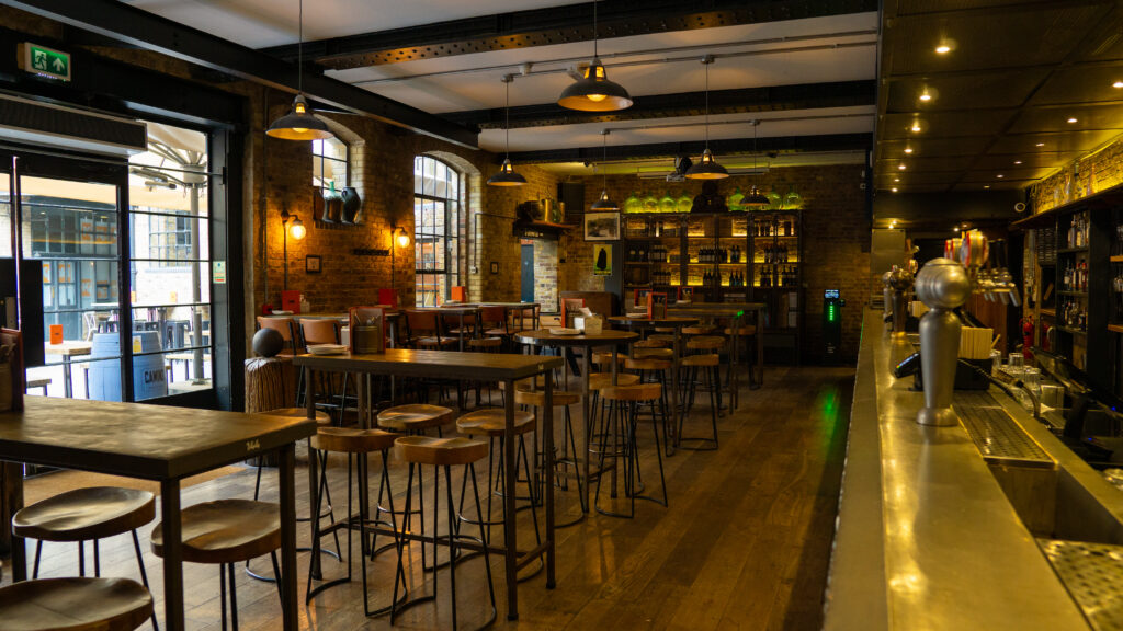 A cozy, dimly lit bar interior with high wooden tables and stools. Exposed brick walls and industrial light fixtures create a warm atmosphere. The bar counter runs along the right side, with shelves of bottles behind it.