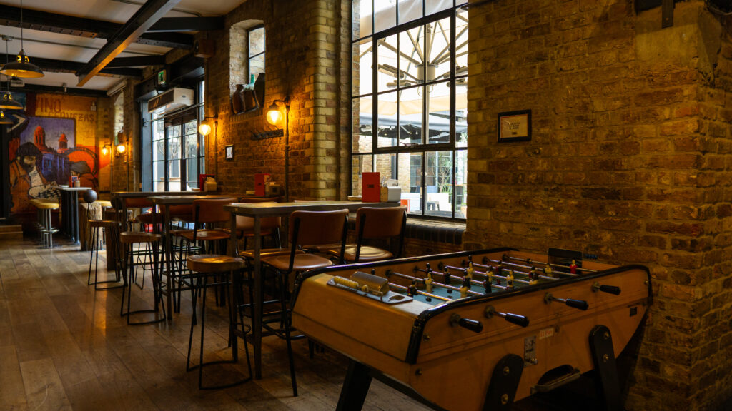 A cozy bar interior with exposed brick walls, wooden floors, and tables with high chairs. A foosball table is in the foreground. Warm lighting and large windows enhance the ambiance. A mural decorates one of the walls.