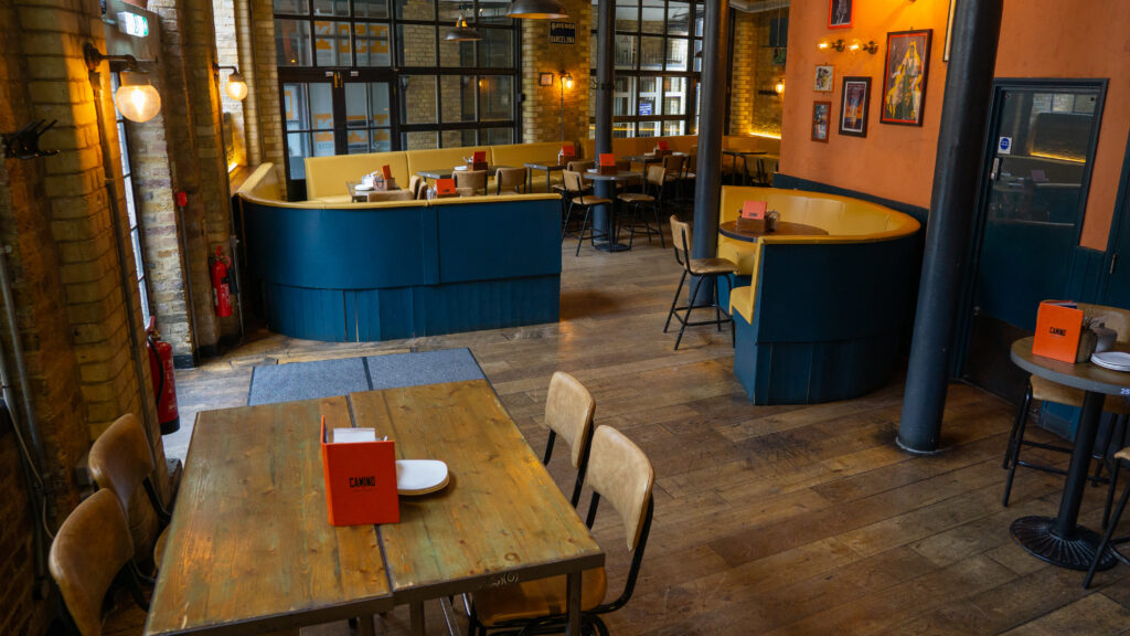 A cozy restaurant interior with wooden tables and chairs. Bright orange menu holders are placed on tables. The space features exposed brick walls, warm lighting, and framed pictures on the walls, creating a welcoming atmosphere.