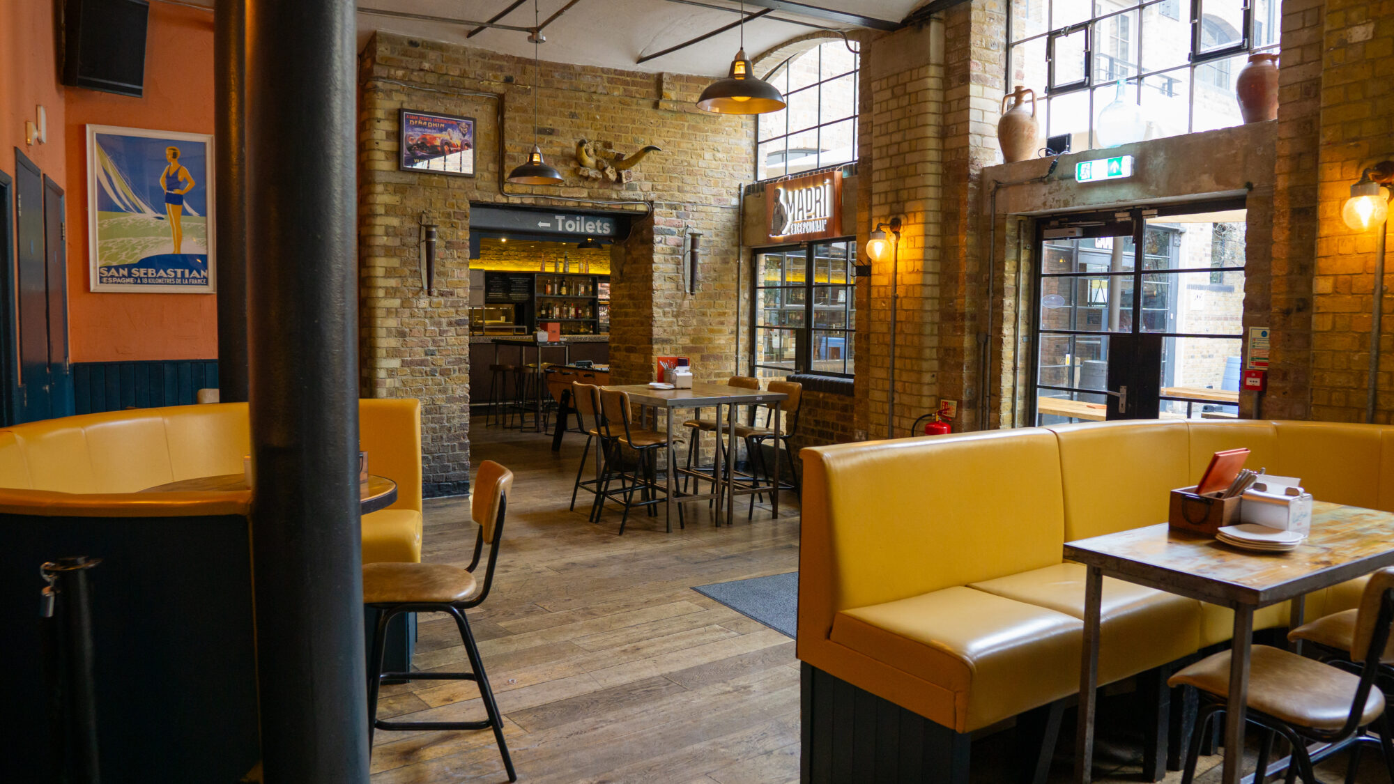 A cozy restaurant interior with yellow booth seating, wooden tables and chairs. Exposed brick walls feature framed artwork. Large windows let in natural light, and a sign for the toilets is visible in the background.