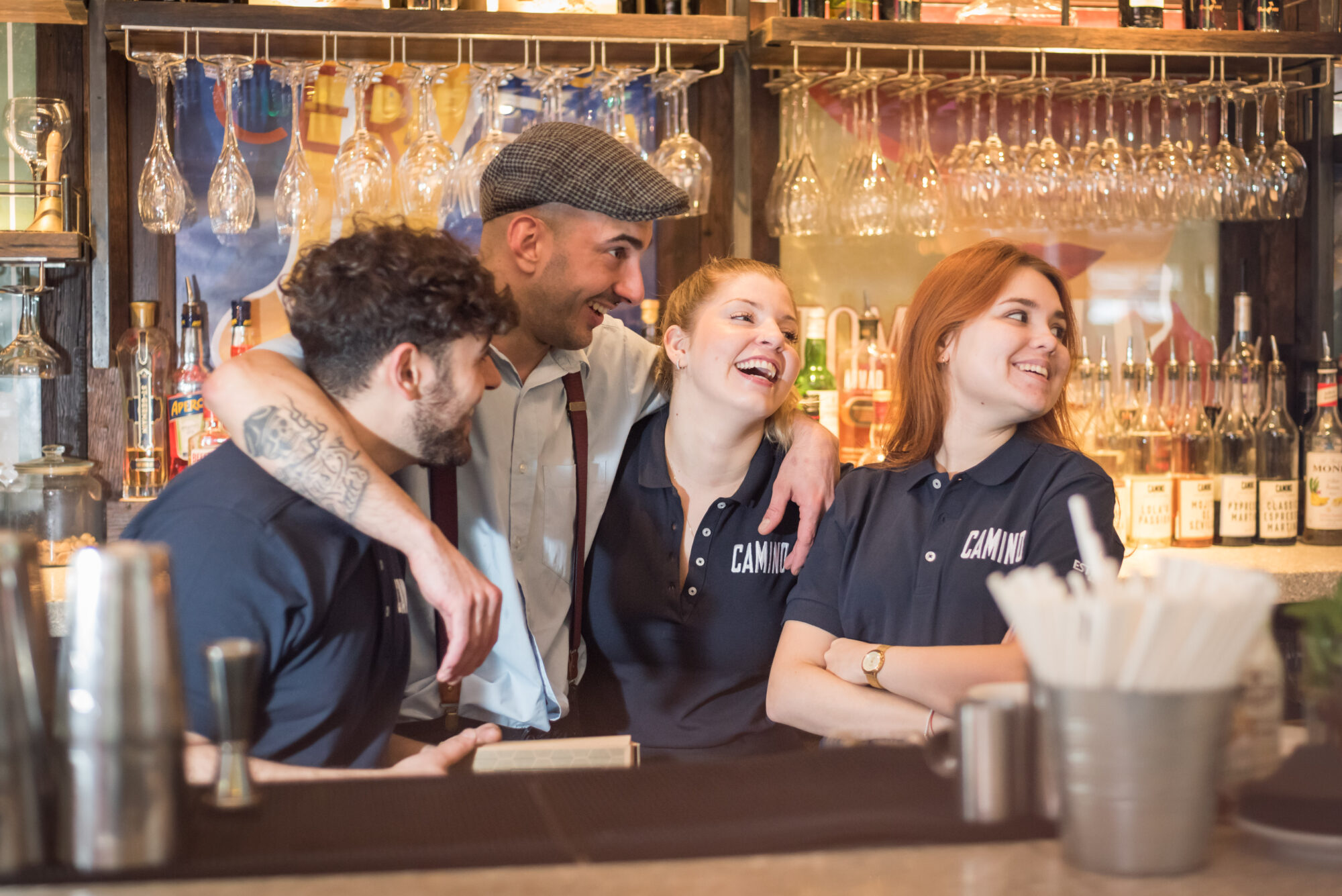 A group of four people, three women and one man, stand behind a bar. They are smiling and interacting happily. The man, wearing a cap, has his arms around them. Shelves with glasses and bottles are in the background.