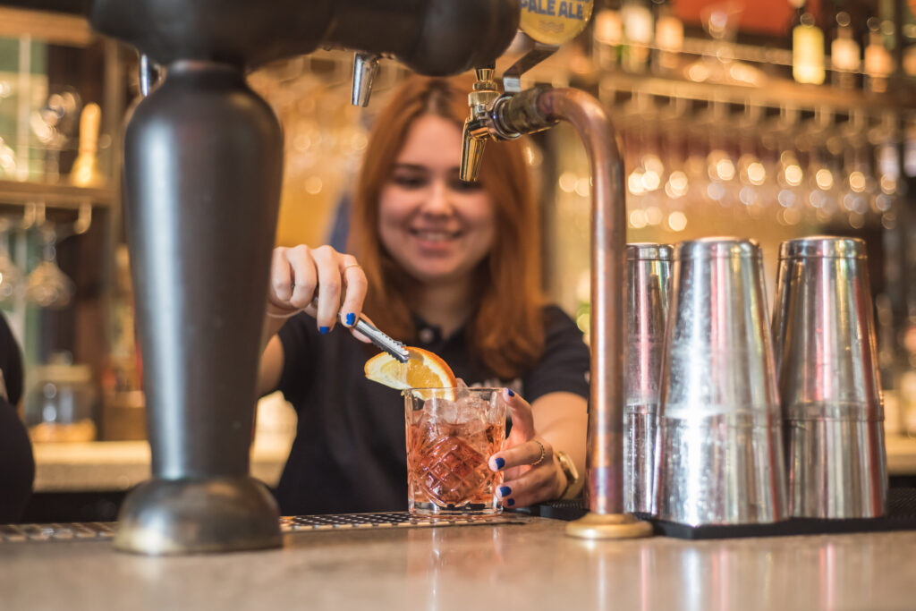 A bartender with brown hair is preparing a cocktail at a bar, adding a slice of orange to a glass with ice. Several metal shakers and beer taps are visible on the counter.