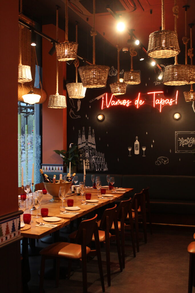 A cozy restaurant interior with a long wooden table set for dining. Wicker lights hang from the ceiling. A neon sign on the wall says 