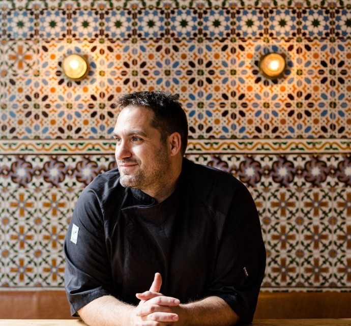 A person in a black chef's coat sits at a table against an ornate, multicolored mosaic tile wall with two circular lights. They look to the side, resting their hands on the table.