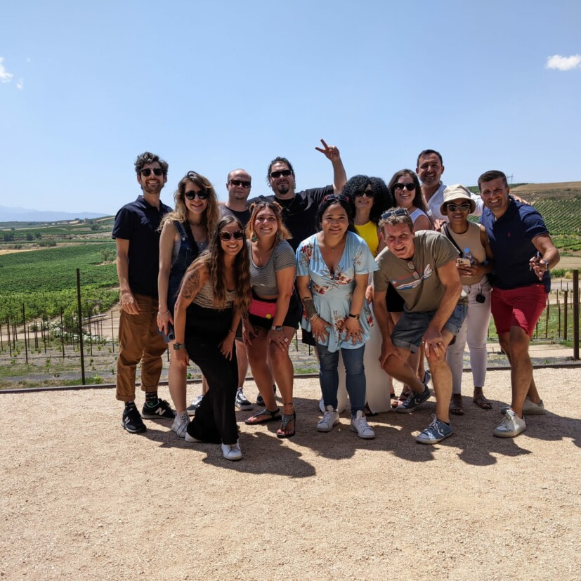 A group of thirteen people posing for a photo in a sunny outdoor setting. They stand on a sandy surface with green vineyards and hills in the background. Most are smiling, and one person is making a peace sign.