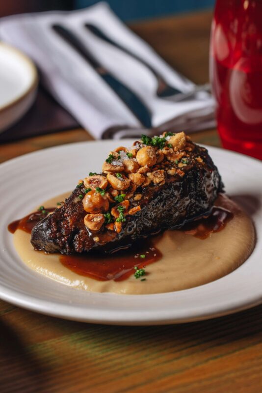 A gourmet dish featuring a seared piece of meat garnished with chopped nuts and herbs, presented on a white plate. The meat rests on a smooth beige puree and is drizzled with a brown sauce. A glass and napkin with cutlery are blurred in the background.