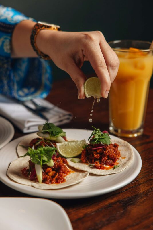 A hand squeezes lime juice over three tacos topped with cilantro and onion on a plate. A glass of orange beverage is in the background, and the table is set with napkins, indicating a cozy dining setting.