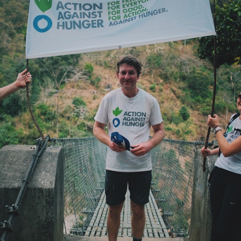 A man in an Action Against Hunger T-shirt stands on a bridge holding a cap, flanked by two people holding a banner with the same logo. The backdrop features a lush, green hillside.