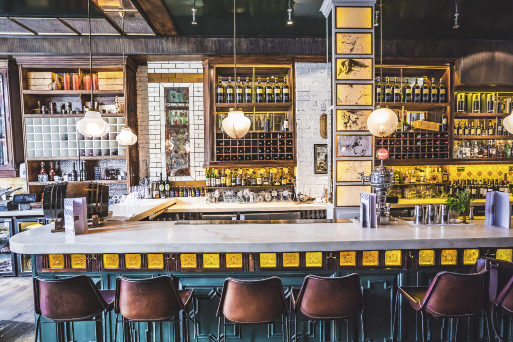 A stylish bar interior with a marble countertop, surrounded by high-backed stools. Shelves behind the bar display bottles and glassware. Warm lighting from pendant lights creates a cozy atmosphere, with wooden accents and decorative tiles.