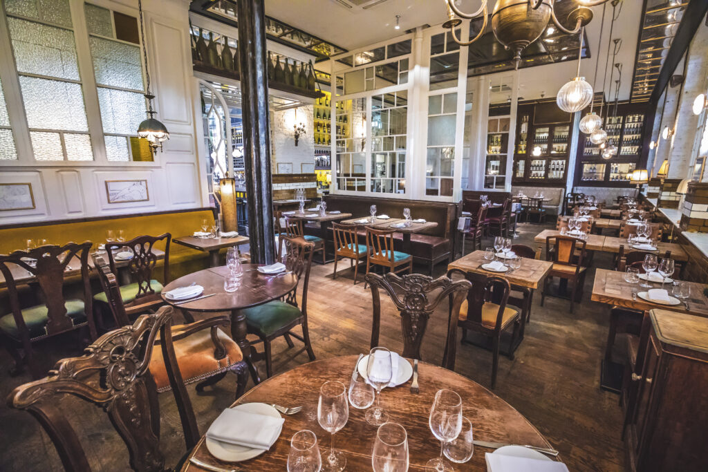 A cozy restaurant interior with wooden tables and chairs, set with plates and glasses. Warm lighting from hanging lamps creates an inviting atmosphere. Decor includes high ceilings, partitioned glass sections, and a mix of vintage and modern elements.