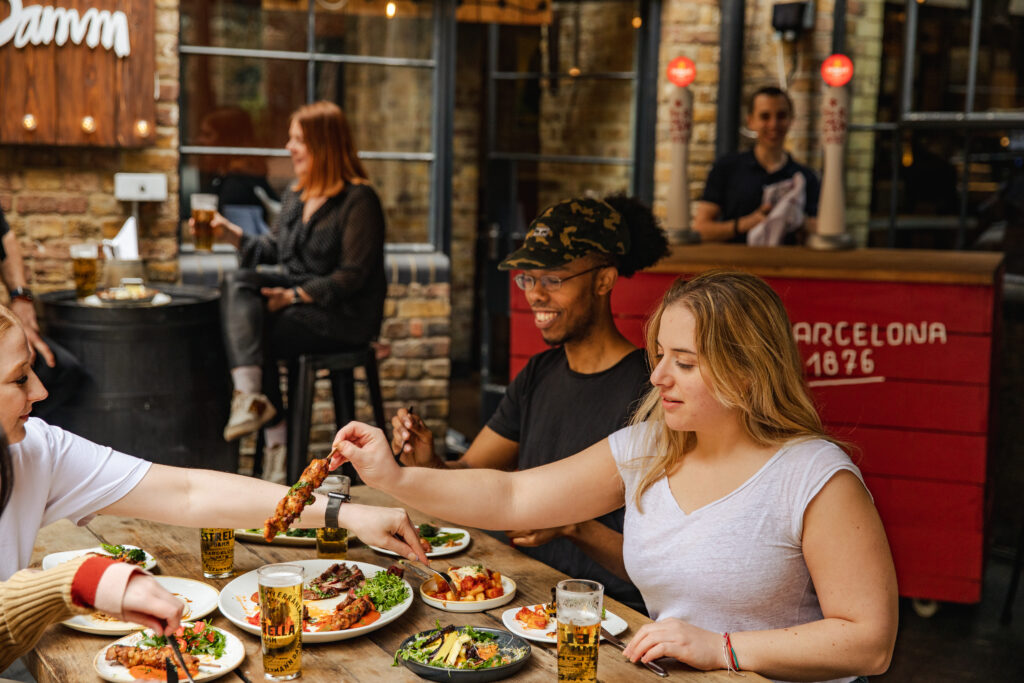 People are enjoying a meal at an outdoor restaurant. They're sharing dishes on a table filled with various foods and drinks. The ambiance is lively, with another person seated at a nearby table and more diners in the background.