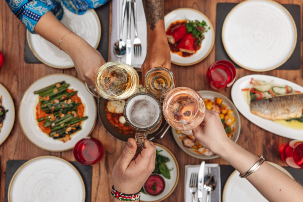 A group of people clinks glasses over a wooden table set with assorted dishes, including a fish platter, green beans, and a vegetable salad. The glasses contain white wine, rosé, and beer. Plates and red glasses surround the meal.