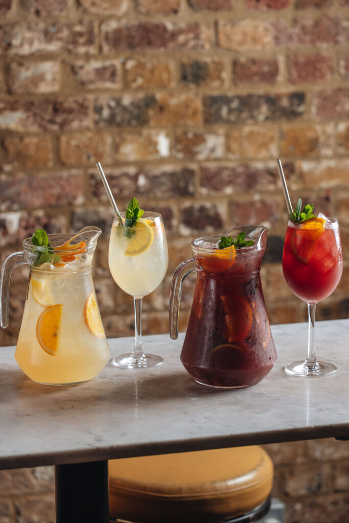 Four drinks on a table: two pitchers of iced tea with citrus and mint, one glass with a lemon slice, and a red drink with an orange slice. Brick wall background.