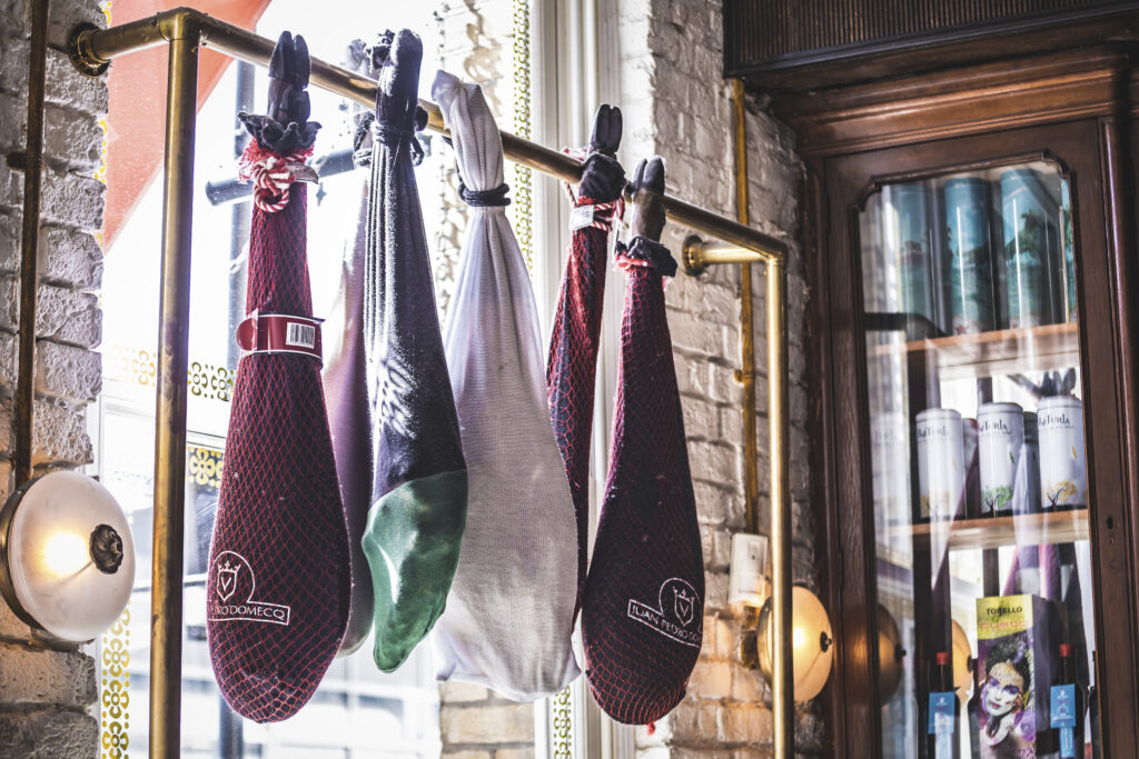 A row of cured meats hangs from a gold-colored bar against a brick wall. Two large haunches are visible, wrapped in red and black coverings. A glass cabinet with items inside is on the right side of the image.