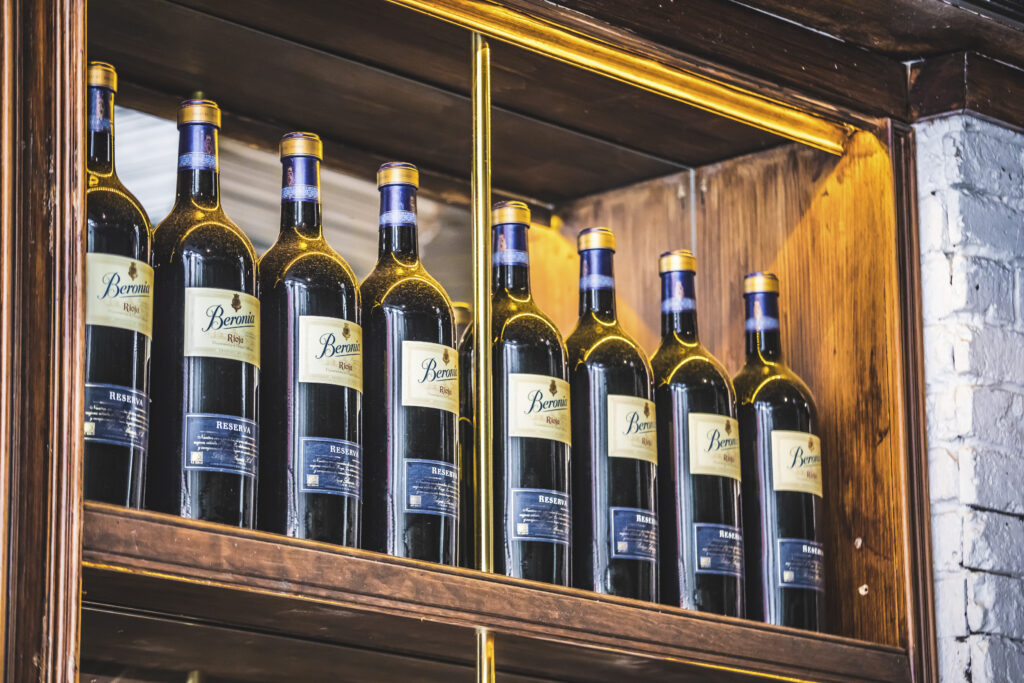 Several wine bottles with blue labels are neatly arranged on wooden shelves with a warm light highlighting them. The shelf is part of a wooden cabinet against a brick wall.