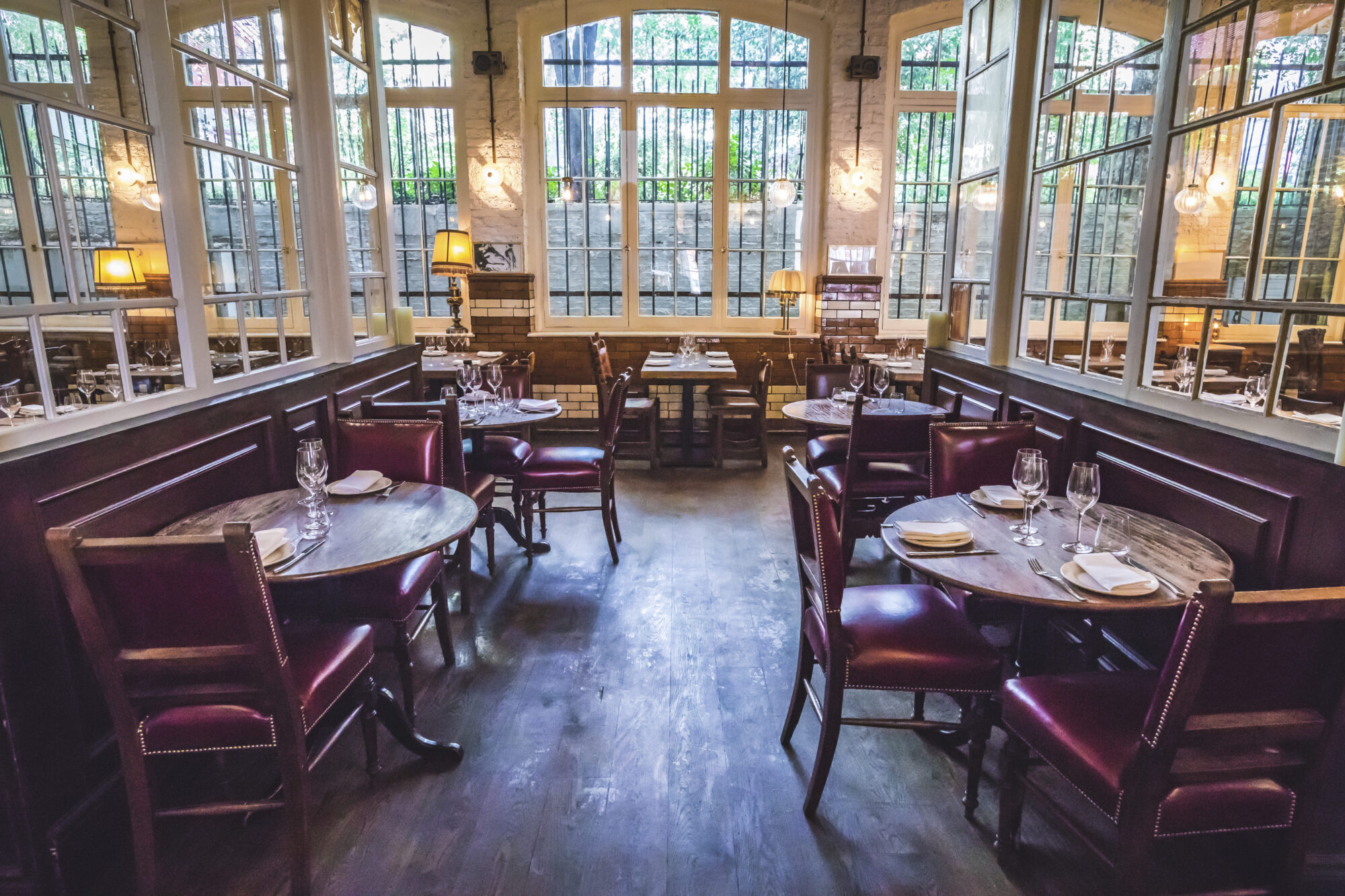 A cozy, elegant restaurant interior with round wooden tables and red leather chairs. Large windows line the walls, letting in natural light. Each table is set with white napkins, cutlery, and wine glasses. Soft, warm lighting enhances the ambiance.